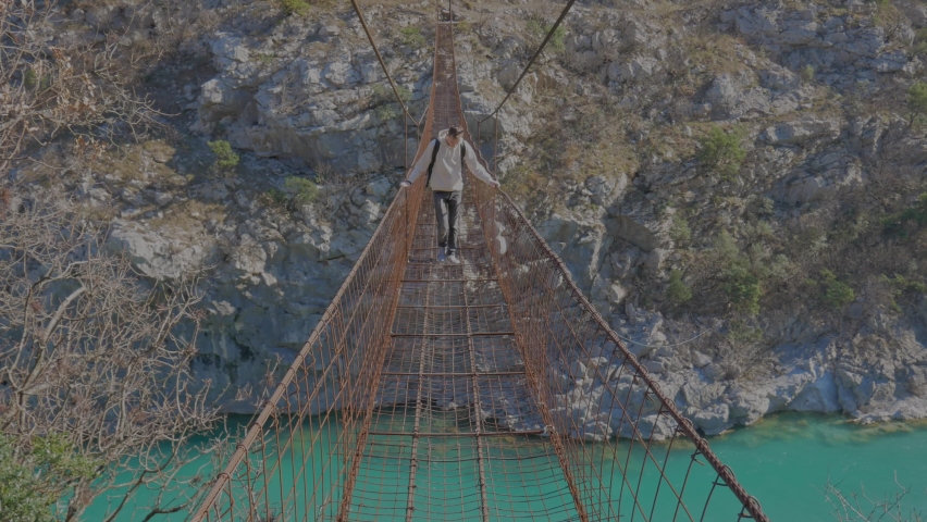 Young millennial man traveler carefully walks by suspension bridge under canyon with river on sunny day. Dangerous walking through the bridge. Monenegro, Podgorica