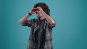 Hipster with his hands in his eyes, spying on his co-workers. Curly-haired teenager. Charismatic hipsterv Studio video, Blue background, Portrait.4k