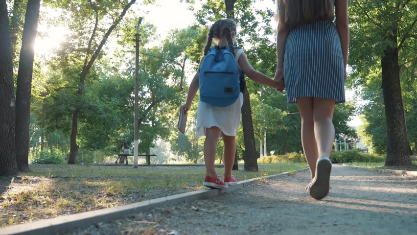 Schoolgirl goes to school with mother. Happy family goes to school in park. Schoolgirl holds her mother hand. Family goes to school. Happy schoolgirl with backpack and book goes through park to school Royalty-Free Stock Footage #1068323201
