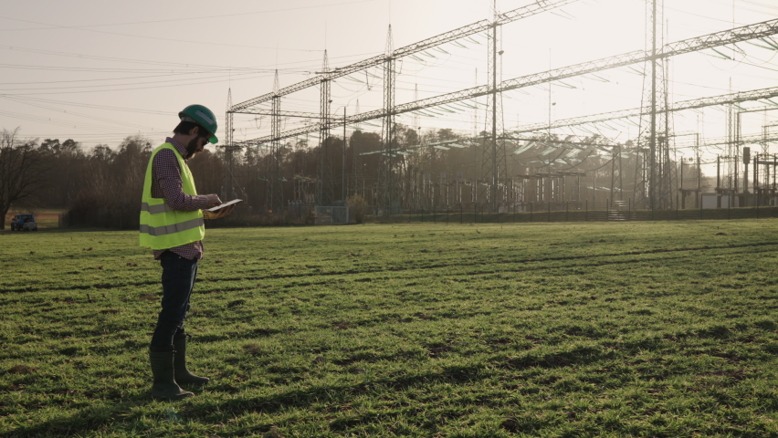 Electrical engineer wearing a helmet and safety vest working with tablet near high voltage electrical lines power station during sunset shot in 4k super slow motion Royalty-Free Stock Footage #1068325175
