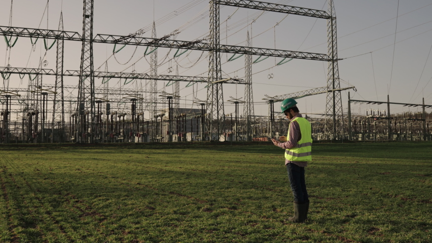 Electrical engineer wearing a helmet and safety vest working with tablet near high voltage electrical lines power station during sunset shot in 4k super slow motion Royalty-Free Stock Footage #1068325178