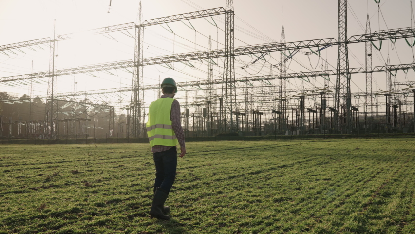 Electrical engineer wearing a helmet and safety vest walking near high voltage electrical lines towards power station during sunset shot in 4k super slow motion | Shutterstock HD Video #1068325202