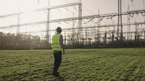 Electrical engineer wearing a helmet and safety vest walking near high voltage electrical lines towards power station during sunset shot in 4k super slow motion