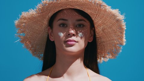 Young brunette asian woman in a straw hat puts some sunscreen on her nose, enjoys the sun and smiles for the camera against blue background | Sunscreen application shot for beauty commercial
