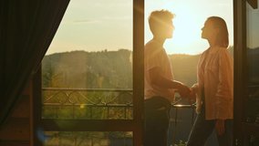 Beautiful young couple standing on the balcony of apartment, talking and kissing on the background of the sunset. Couple of tourists kissing on the balcony of a country house in mountains sunlight.