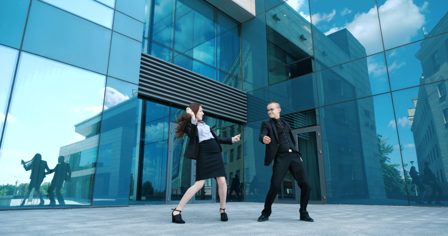 Pair of colleagues woman and man wearing business black suits, dancing together moving their arms and legs on street in open air, two business people having fun after hard day celebrating victory