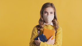 Tired blond teenage student girl wearing protective mask standing with books and intently looking in camera over yellow background. Safety first