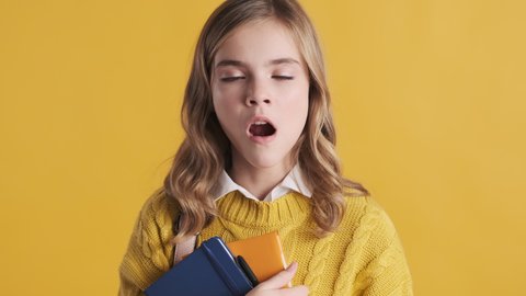Sleepy blond teenage student girl yawning on camera wake up early because of school over yellow background. Study concept