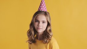 Pretty blond teenage girl wearing party hat celebrating her birthday with friends looking happy over yellow background. Birthday girl