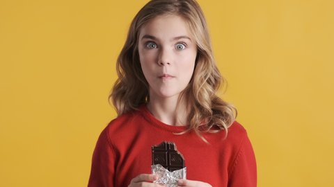 Beautiful blond teenage girl eating chocolate bar waving yes keeping thumb up isolated on yellow background. Sweet tooth girl. Like it gesture