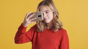 Pretty blond teenage girl in red sweater looking happy using smartphone over yellow background. Attractive girl turning on music in phone and dancing
