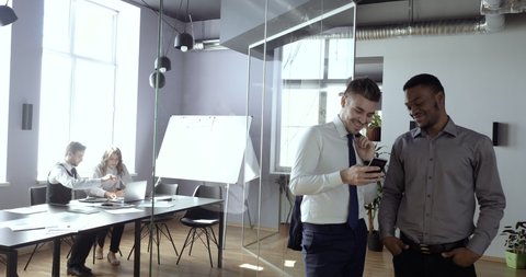 Two business colleagues talking at work relaxing looking at smartphone screen, background of managers routine. African american worker and caucasian man in office using phone to search net together