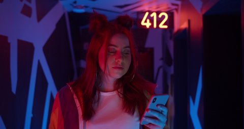 Lviv, Ukraine - May 21, 2020: female person using her mobilephone. Good looking millennilal woman with trendy hairdo looking at phone screen while walking in corridor with neon lights.