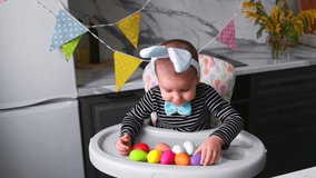 Funny Blond Boy Kid 2 y.o. with Rabbit Ears plays with colored Easter Eggs sitting at the Kitchen table. Holidays Concept.