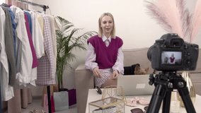 A female stylist blogger talks in front of the camera about glasses of different shapes. A female blogger. Next to the clothes rack. On the couch is a small Pomeranian dog in clothes.