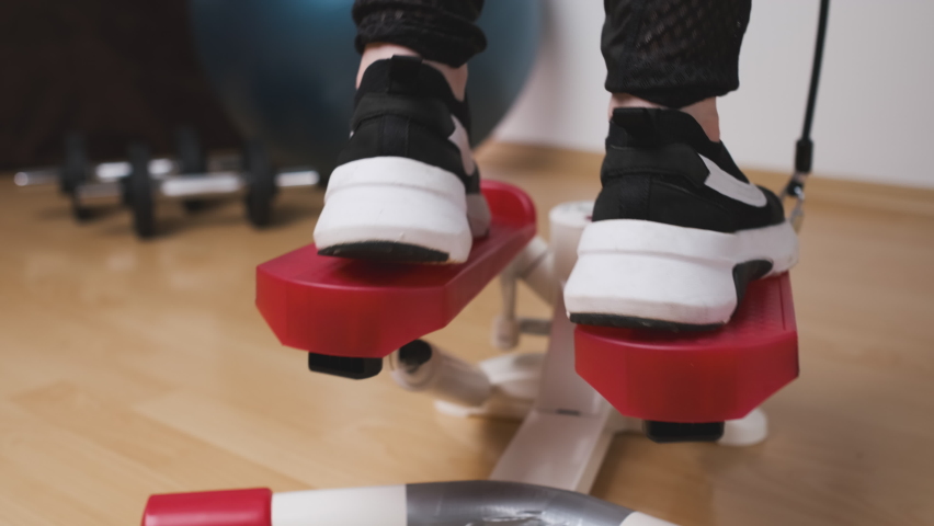 Back view of female legs doing exercises on the twist stepper in sneakers and sportswear at home during lockdown. Workout at home 