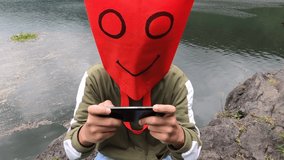 Adorable ugly funny red bag head smiley face enjoying play online game on smartphone seriously while sitting on rock at edge of exotic tropical fresh water lake. Handheld shoot cloudy morning day.
 
