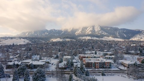 Aerial droneing backward over University of Colorado Boulder campus covered in snow on a winter morning with the mountains of the front range covered in clouds in the background