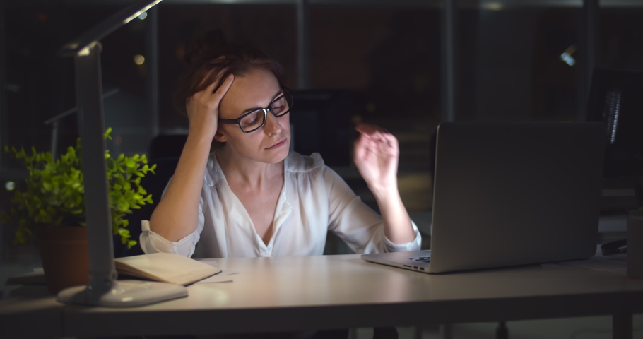 Exhausted young woman at office desk working late at night. Portrait of tired and stressed businesswoman having migraine overworking in office late in evening | Shutterstock HD Video #1068358445