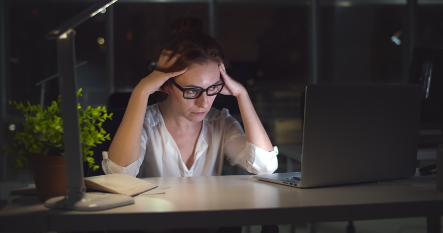 Exhausted young woman at office desk working late at night. Portrait of tired and stressed businesswoman having migraine overworking in office late in evening | Shutterstock HD Video #1068358445