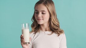 Beautiful blond teenage girl holding glass of milk and smiling in camera over blue background