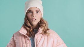Attractive blond teenage girl dressed in pink down jacket and hat trying afraid friend looking fun isolated on blue background