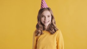 Pretty cheerful blond teenager girl wearing party hat dancing on her birthday over yellow background. Fooling around expression