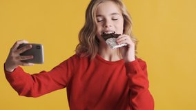 Pretty blond teenage girl taking selfie with chocolate and choosing it for social network over yellow background. Modern technology concept
