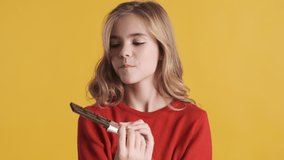 Attractive blond teenage girl looking happy eating chocolate over yellow background