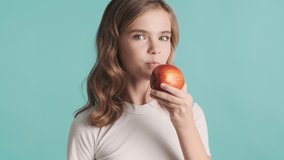 Pretty funny blond teenager girl with wavy hair eating delicious apple and smiling isolated on blue background. Time for snack
