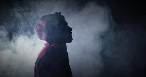 A Handsome Caucasian bearded sexy boy staying in dark black room with white smoke around. A mental sportsman in red hoody staying before competition, thinking and looking up on black background