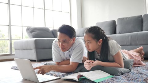 Asian family Father teaches daughter books by using laptop computer lying on the floor in the living room. Happy asian studying at home, study online