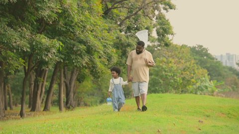Happy african child boy holding box and dad holding dragonfly catcher net walking in park and outdoor. African american man and son holding hands talking and walking to catching dragonflies in garden.