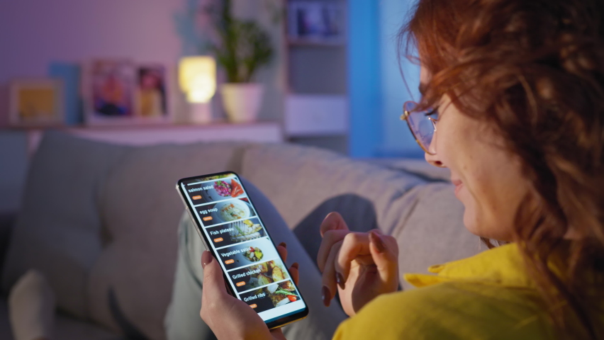 Food delivery, young girl in glasses with mobile phone in hands chooses fast food in an online store while relaxing on sofa in living room in evening | Shutterstock HD Video #1068377897