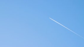 An airplane flies high above the ground leaving an even white trail. Blue sky, natural landscape illuminated by the sun. Beautiful background with clear weather and copy space. Time-lapse 4K video.