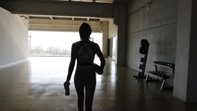Tracking shot of caucasian slim Woman from the back, with a fitness mat walks through an empty gymnasium fitness center to start the exercise routine. Big windows, Silhouette, back lit.