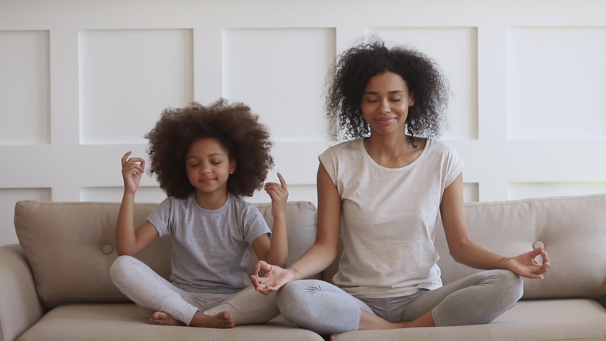 Funny african family young healthy mom teaching cute little kid daughter meditate together sit on sofa at home, calm mindful black mother and child girl do yoga exercise relax together in lotus pose Royalty-Free Stock Footage #1068386123