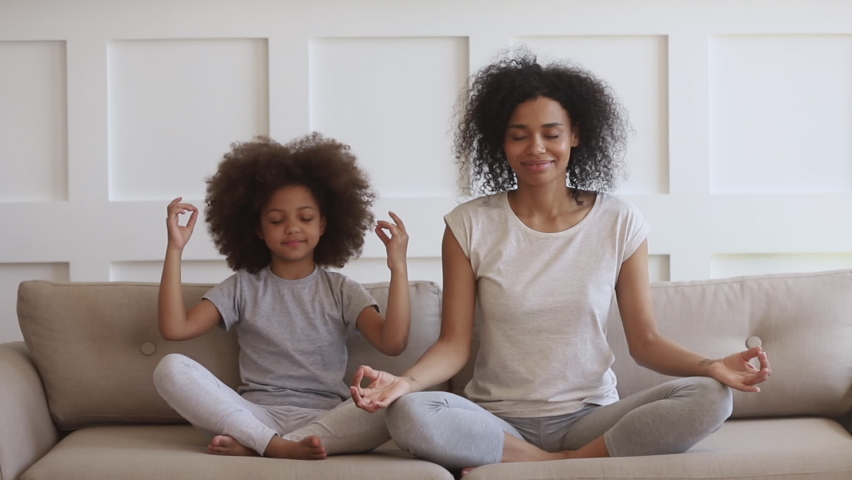 Funny african family young healthy mom teaching cute little kid daughter meditate together sit on sofa at home, calm mindful black mother and child girl do yoga exercise relax together in lotus pose Royalty-Free Stock Footage #1068386123