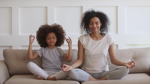 Funny african family young healthy mom teaching cute little kid daughter meditate together sit on sofa at home, calm mindful black mother and child girl do yoga exercise relax together in lotus pose