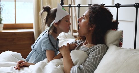 Funny little kid girl wear nurse hat holding stethoscope pretending doctor check mother throat in bedroom, cute preschool child daughter looking inside open mouth having fun playing with mum in bed