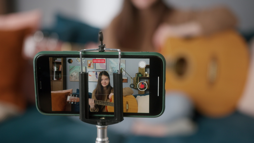 Music blogger is playing guitar, shooting herself on video. Musician is making vlog. Woman is teaching how to play guitar. Student is playing acoustic instrument in front of camera. 
