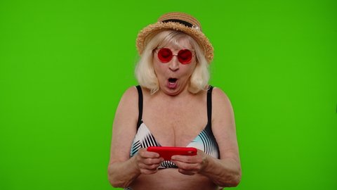 Senior woman tourist in swimsuit bra playing a video game on a mobile cell phone, using mobile app drive simulator, spending leisure time on internet entertainment on chroma key background. Exited granny.