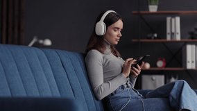 Portrait of smiling young girl wearing headphones, choosing favorite music tracks in mobile application,texting messages, browsing internet, watching video, playing online game on smartphone at home.