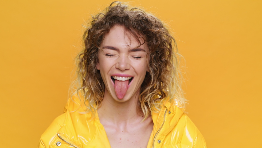 Portrait Happy Young Woman Smiles Coquettishly Bites her Lips Sticks out Tongue Looks at Camera. Glamorous Girl Curly Hair in Yellow Jacket on Yellow Background. Fashion. Monotone. Positive Emotions Royalty-Free Stock Footage #1068406853