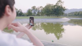 Happy young man holding a smartphone, looking at the screen, talking on a video call while sitting on the grass in the park, online technology, mobile application, social distancing, self isolation
