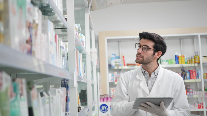 Male pharmacist using digital tablet and wear gloves reduce exposure checking stock inventory in modern pharmacy. Prevention of infectious diseases, touch, New Normal Concept  Royalty-Free Stock Footage #1068415970
