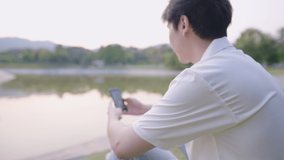 Asian lonely man video calling to his friend while sitting next to pond park with raising two fingers and waving hands to phone, people and distant relationship, green environment, self isolation 