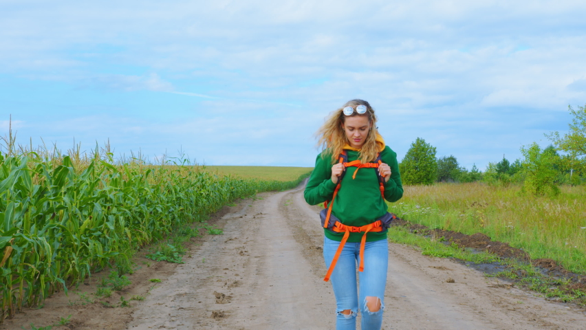 A woman goes along a cornfield on a dirt road. A hitchhiker traveler puts on sunglasses while moving forward