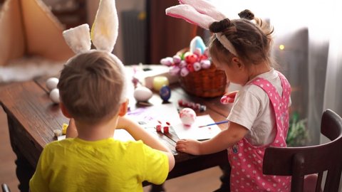 Brother and sister are painting Easter eggs for Easter. Happy children paint eggs in a playful way. Children have rabbit ears on their heads