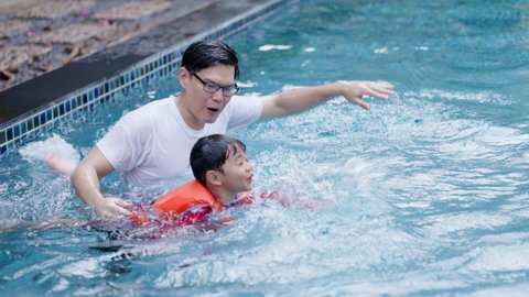 Asian family father is teaching and playing with his son swimming in pool. Swimming teacher training student to wear swimming life jacket. Happy holidays of summer. Concept Workout